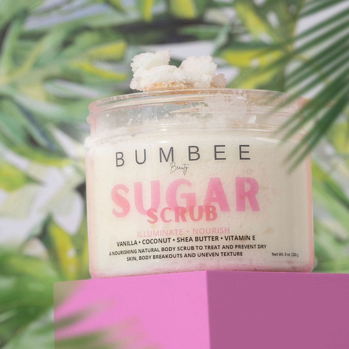 A jar of sugar scrub placed in a tropical setting, with the scrub's textured surface visible on top of the jar. The image evokes a sense of relaxation and indulgence, highlighting the product's natural ingredients and luxurious feel
